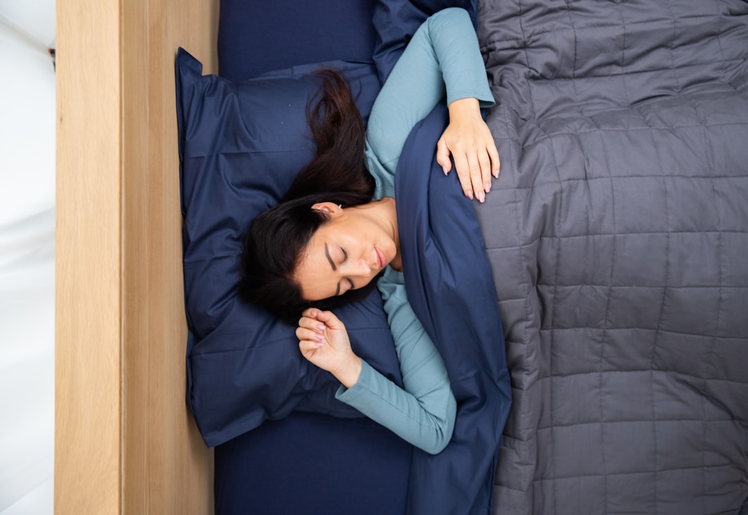 A woman with long dark hair is sleeping peacefully in a bed. She is lying on her side, covered with a dark grey blanket, using a blue pillow. She is wearing a light blue long-sleeve shirt and her right arm is resting on the blanket.