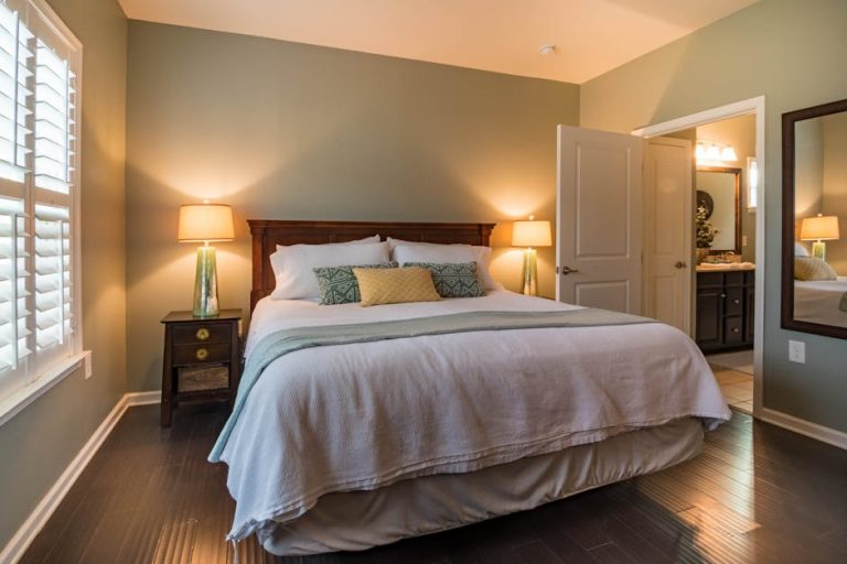 A cozy bedroom with a large bed adorned with white and green bedding. Two bedside tables with lamps flank the bed. There's a mirror on the right wall, a window with white shutters on the left, and an open door leading to a bathroom in the background.