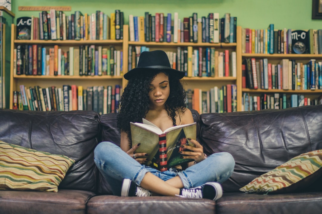 A person with curly hair wearing a black hat sits cross-legged on a dark brown leather couch, reading a book. The background is filled with bookshelves containing various books. The couch has several patterned cushions.