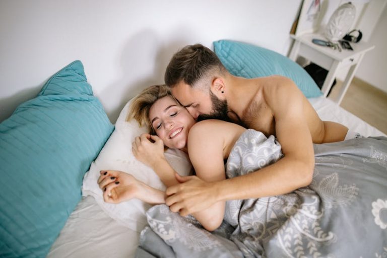 A shirtless couple is lying in bed, partially covered by a grey and white patterned blanket. The man is kissing the woman's cheek while she smiles with her eyes closed. The bed has blue pillows and a white nightstand is visible in the background.