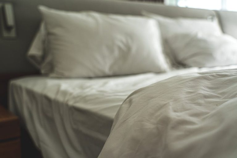 A bed with neatly arranged white sheets and pillows. The bed is made and looks clean and inviting. The headboard is visible in the background, and a glimpse of a side table can be seen on the left side of the image.