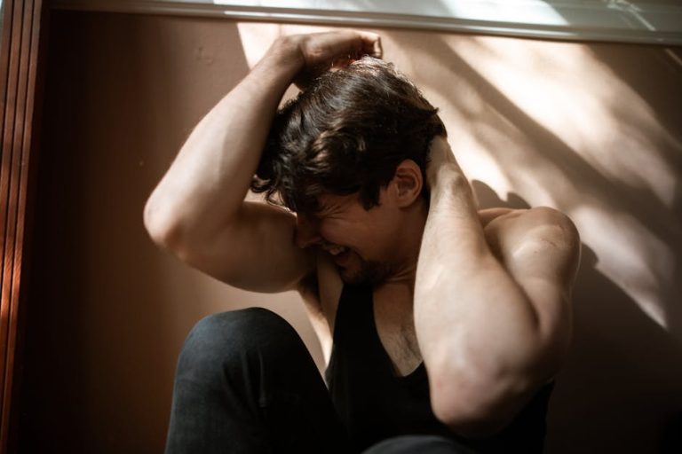 A man with dark hair and a beard sits on the floor, wearing a black sleeveless top. He is gripping his head and neck with both hands, and has a pained or distressed expression on his face. Sunlight streams through blinds, casting shadows on the wall behind him.