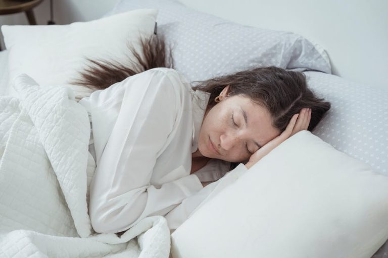 A person with long dark hair is sleeping peacefully in bed. They are lying on their side, head resting on one hand, covered by a white quilt. The bed is adorned with white pillows and light grey sheets with white polka dots.