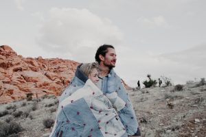 A couple stands together wrapped in a quilted blanket in a rocky desert landscape. The sky is cloudy, and they appear to be enjoying the view. Red rock formations and sparse vegetation surround them.