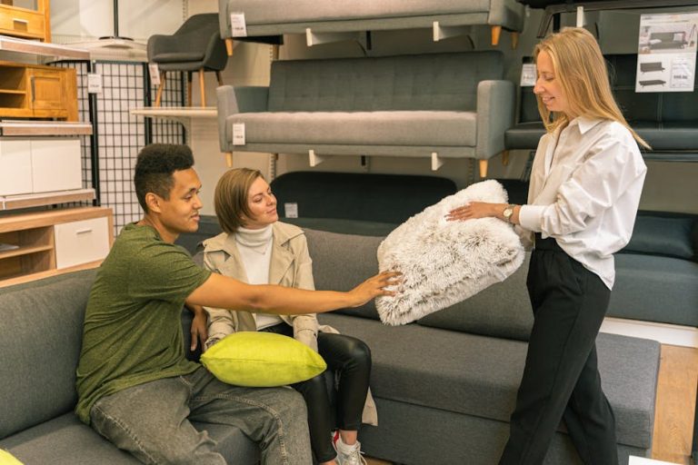 Three people in a furniture store: a man in a green shirt and a woman in a beige trench coat sit on a gray couch, while a woman in a white blouse shows them a fluffy white cushion. They are surrounded by various pieces of furniture on display.