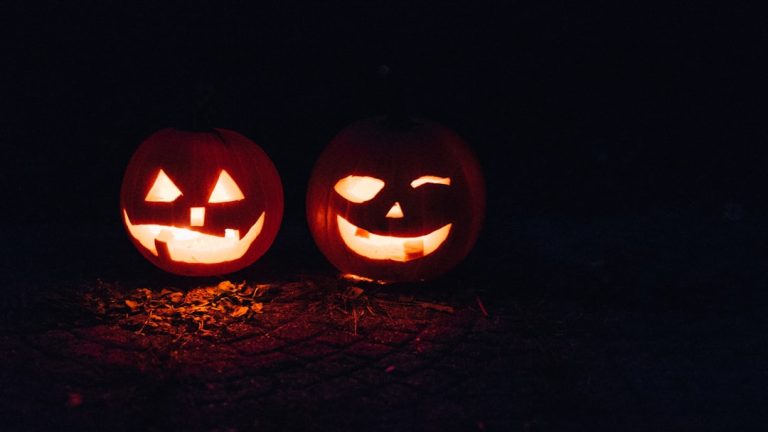 Two carved pumpkins glow with candlelight in a dark setting. The pumpkin on the left has a classic jack-o'-lantern design with triangle eyes and a big, toothy smile. The pumpkin on the right has one eye winking and a lopsided grin.