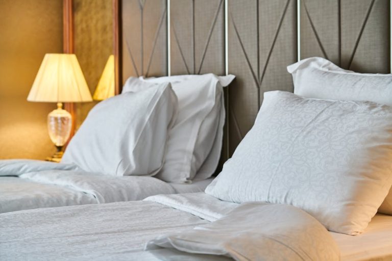 A cozy bedroom features two neatly made twin beds with white bedding. Each bed is adorned with two pillows, and a stylish table lamp with a beige shade sits on a nightstand between them, casting a warm glow in the room. The headboard has a geometric design.