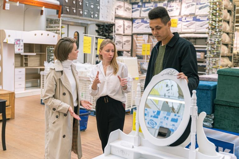 Three people are in a store, possibly discussing or shopping for furniture. One woman in a beige trench coat is speaking to a woman in a white blouse and black pants. A man in a black jacket stands nearby, looking at a white vanity mirror on display.