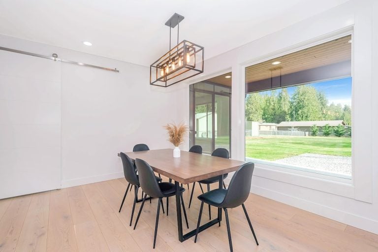 A modern dining room with a wooden table and six black chairs. A contemporary rectangular chandelier hangs above the table. The large window provides a view of a green yard and trees. The room has light wood flooring and white walls.