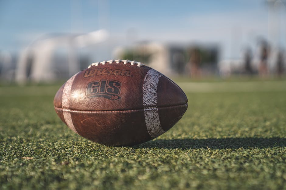 Close-up of a worn-out Wilson football lying on a grassy field. The background is blurred, showing an outdoor sports area, possibly a football field, with vague outlines of equipment and structures under a clear sky.