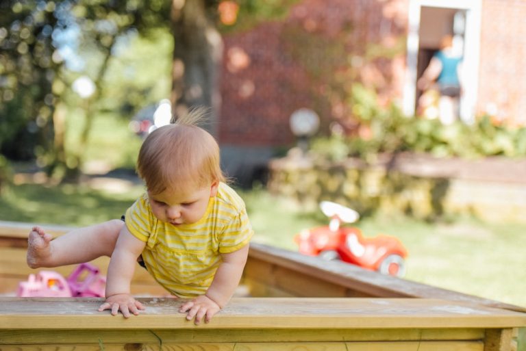 A baby in a yellow striped onesie is climbing over the edge of a wooden sandbox outdoors. A red toy car is on the grass nearby, and a person is visible in the background near a brick building. It's a sunny day, with trees and greenery around.