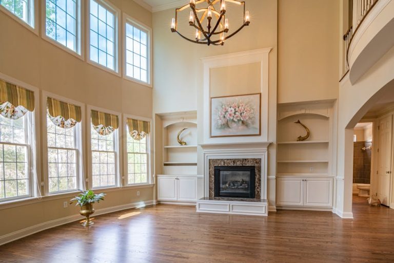 A spacious living room with tall windows, a chandelier, and hardwood floors. The high ceiling features a large hanging light fixture. The room has built-in shelves, a fireplace with a stone surround and artwork above it, and a plant by the windows.