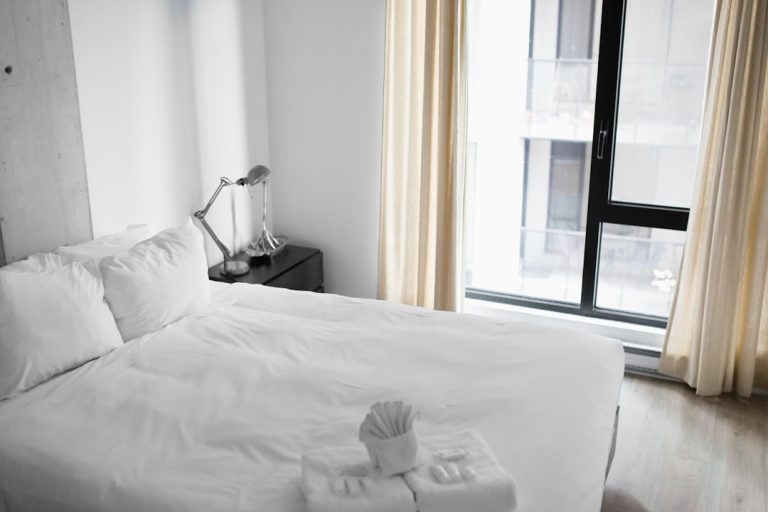 A minimalistic bedroom features a neatly made bed with white linens, a folded towel, and toiletries arranged on it. Next to the bed is a nightstand with a modern desk lamp. The room is softly lit by natural light filtering through beige curtains over large windows.