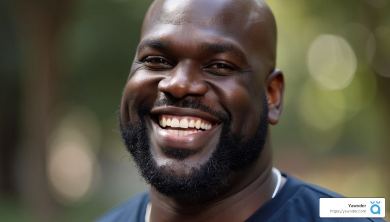 A man with a beard is smiling broadly against a blurred, natural outdoor background. He is wearing a dark shirt. In the bottom right corner, there is a logo featuring the word "Yawner" and a small icon.