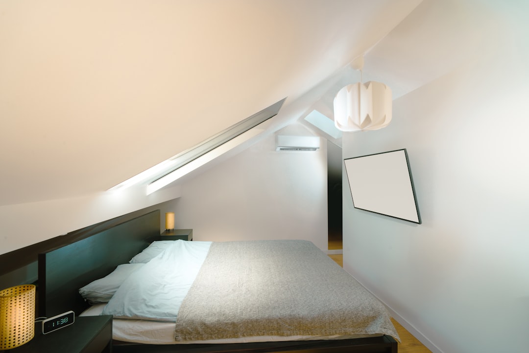 A minimalist attic bedroom featuring a large bed with grey bedding, a slanted roof with a skylight, a wall-mounted TV, and modern pendant lighting. Nightstands with lamps are on each side of the bed, and there is light wood flooring.