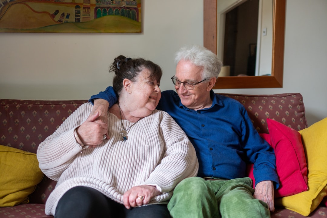 An elderly couple sits on a sofa, smiling and embracing. The woman is wearing a light pink sweater, and the man is in a blue shirt and green pants. They are surrounded by colorful pillows and have a painting on the wall behind them.