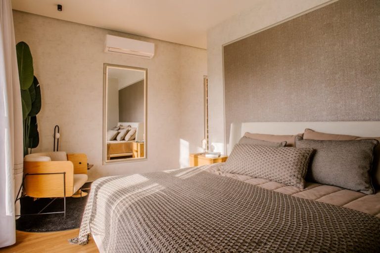 A cozy, modern bedroom with a neatly made bed featuring neutral-toned bedding, a large textured headboard, and a mirror on the wall. A stylish wooden chair and a dark rug are near a leafy plant and a window, letting in natural light. An air conditioner is mounted above.