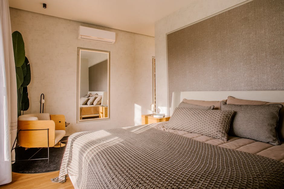 A cozy, modern bedroom with a neatly made bed featuring neutral-toned bedding, a large textured headboard, and a mirror on the wall. A stylish wooden chair and a dark rug are near a leafy plant and a window, letting in natural light. An air conditioner is mounted above.