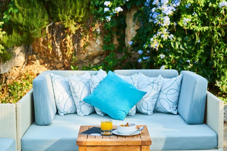 A cozy outdoor seating area features a light gray couch adorned with patterned white and gray pillows and a bright blue accent pillow. In front, a wooden table holds a glass of orange juice, a plate with snacks, and a napkin. Lush greenery and flowers surround the space.