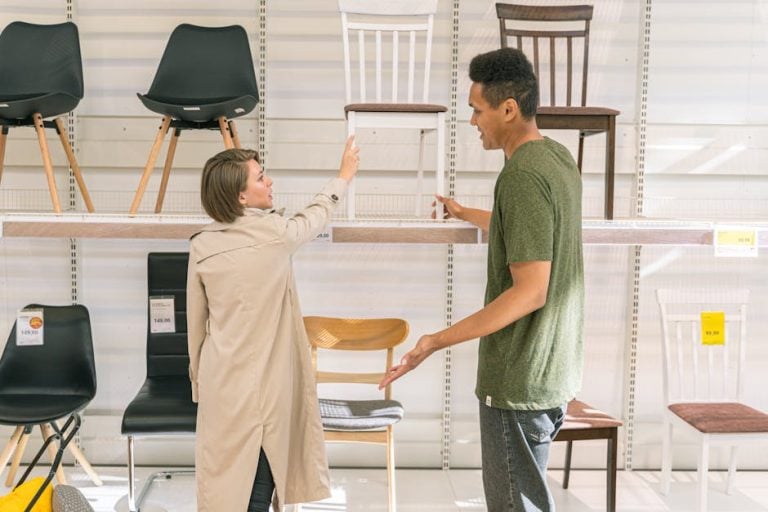 A woman and a man are shopping for chairs in a furniture store. The woman in a beige coat points toward a row of chairs on display, while the man in a green shirt looks on and gestures with his hands. Various chair styles and price tags are visible on the shelving.