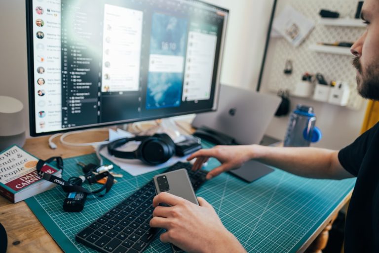 A person uses a smartphone and keyboard while working at a cluttered desk with a large monitor displaying various chat windows. The desk also includes a laptop, headphones, a water bottle, and stationery items, creating a busy workspace.