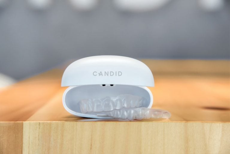 A white orthodontic aligner case labeled "CANDID" is open on a wooden surface, revealing clear aligners partially protruding from the case. The background is blurred, emphasizing the focus on the aligner case and aligners.