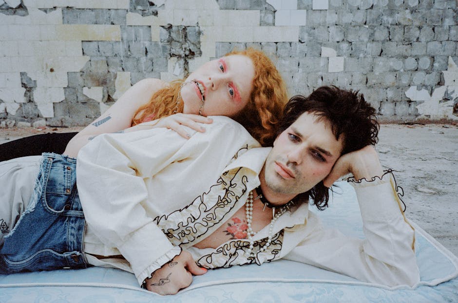 A person with long red hair wearing a black top and a person with dark hair wearing a white shirt with ruffles are lying on a mattress outdoors, against a dilapidated wall. The red-haired individual leans on the other person, who looks away, deep in thought.