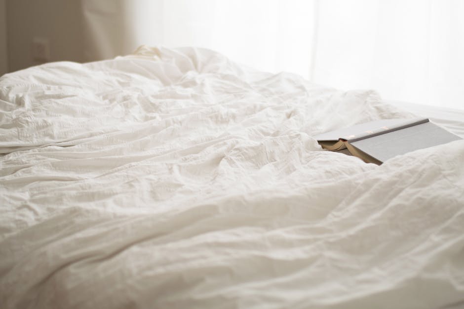 A cozy, unmade bed with white sheets and a closed book resting on top. Soft natural light filters through a sheer curtain in the background, creating a serene and inviting atmosphere.