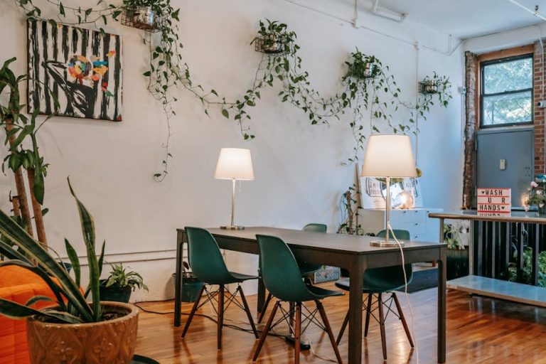 A cozy room with a wooden table and green chairs, adorned with two table lamps. Plants hang on the white walls, and a bright orange sofa is partially visible. A window and a sign reading "Wash Your Hands" complete the decor.