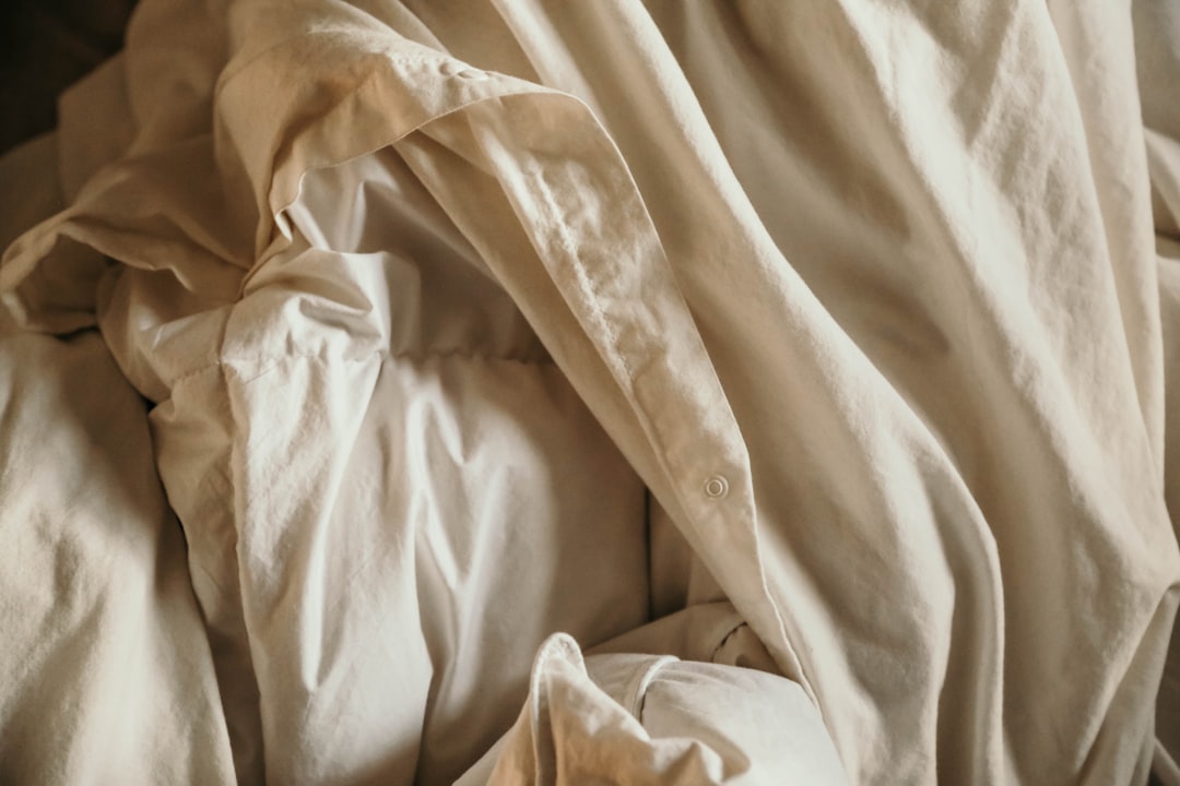 Crumpled, off-white bed sheets create an abstract pattern. The fabric appears soft and well-worn, with natural folds that catch the light, creating gentle shadows.