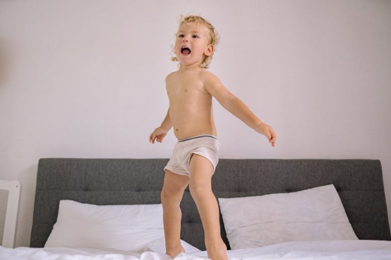 A cheerful toddler with curly hair jumps on a bed, wearing only light-colored shorts. The bed is made with white linens and has a gray headboard. The child's expression is energetic and joyful, and the background is a plain white wall.