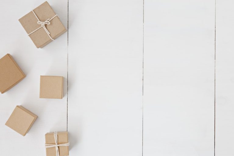 Five small brown gift boxes tied with string are scattered on a white wooden surface. The boxes vary in size and are arranged mostly on the left side of the image, leaving a large empty area on the right.