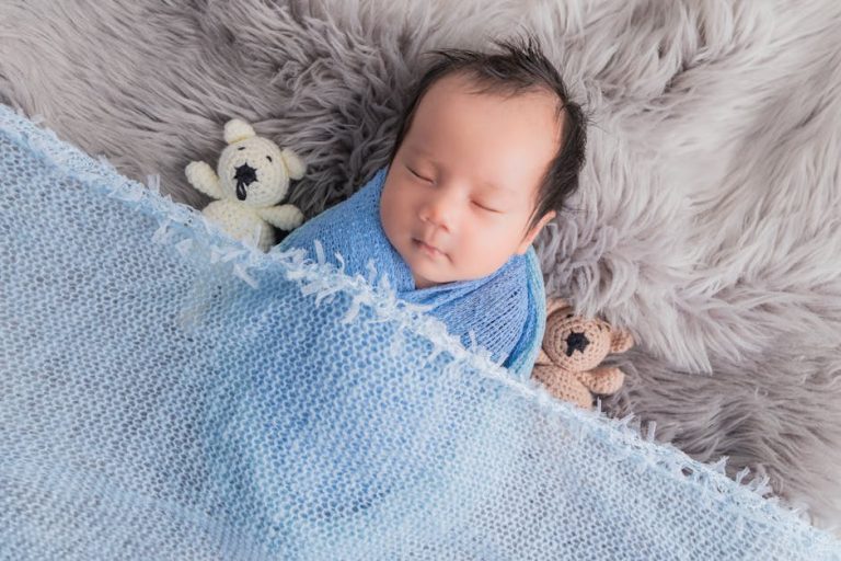 A baby peacefully sleeps swaddled in a blue blanket, surrounded by two small knitted teddy bears on a soft gray furry surface.