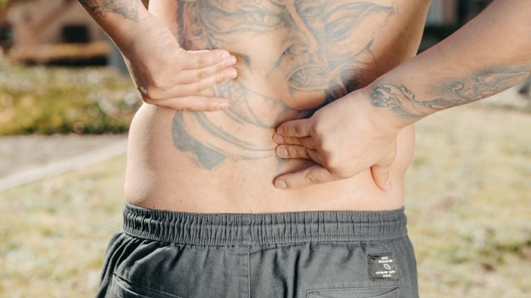 A person stands outdoors, showing their back with detailed tattoos. They are wearing dark shorts and using their hands to massage their lower back. A grassy area is visible in the background.