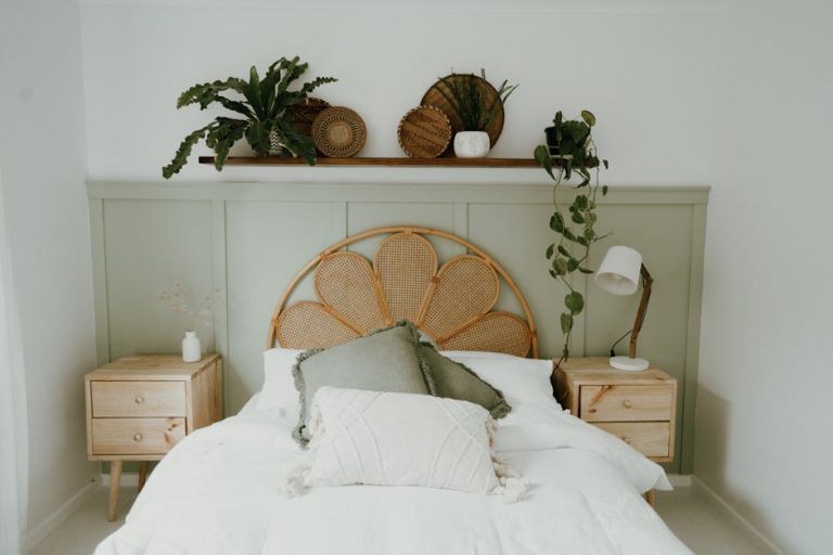 A cozy bedroom features a bed with a rattan headboard and white bedding. There are light wooden nightstands on each side, a green accent wall with a shelf above holding plants and decorative items. A reading lamp is on the right nightstand.