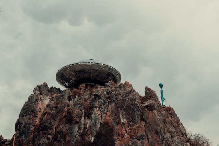 A UFO-shaped structure sits atop rocky cliffs under a cloudy sky, with a small green alien figure standing nearby.