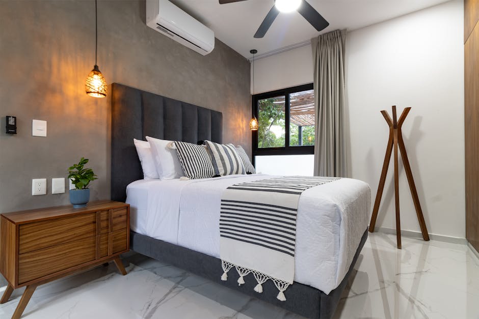 A modern bedroom with a large bed, striped throw blanket, and pillows. Wooden nightstand with a plant and hanging pendant lights. Gray accent wall and window with curtains. Air conditioner and ceiling fan visible. Marble-tiled floor.