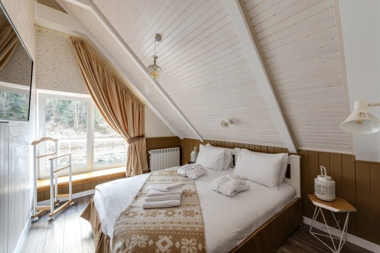 A cozy attic bedroom with a sloped wooden ceiling, featuring a large bed with white and patterned bedding. Natural light streams through a window with drawn curtains, and there's a small wooden side table with a lamp.