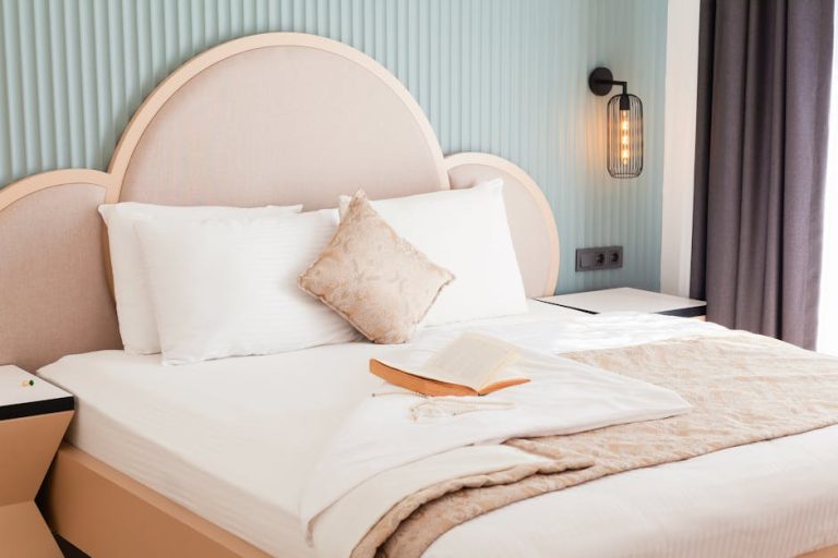 A neatly made bed with a light beige headboard and white bedding. A decorative pillow and an open book rest on the bed. The wall is a soft turquoise, and a modern wall sconce provides lighting. A curtain hangs on the right side.