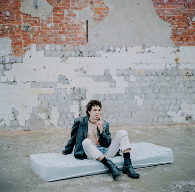 A person with curly hair sits on a mattress in an industrial space. They are wearing a dark jacket, light pants, and boots. The background features exposed brick and peeling plaster.