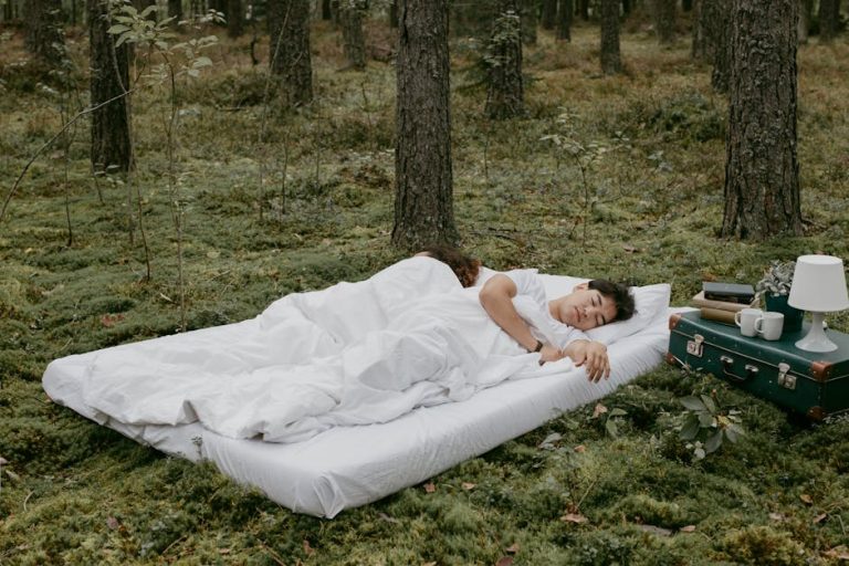 A person sleeping on a white mattress placed on a forest floor covered with green moss. Beside them is a vintage suitcase topped with a lamp, a cup, a teapot, and a dish, creating a cozy outdoor setting.