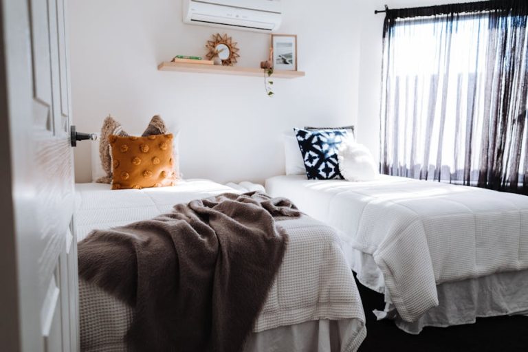 A cozy bedroom with two neatly made twin beds, each adorned with decorative pillows. A soft brown blanket is draped over one bed. Above the beds, a shelf holds a small plant and a framed picture. Sunlight streams through sheer curtains.