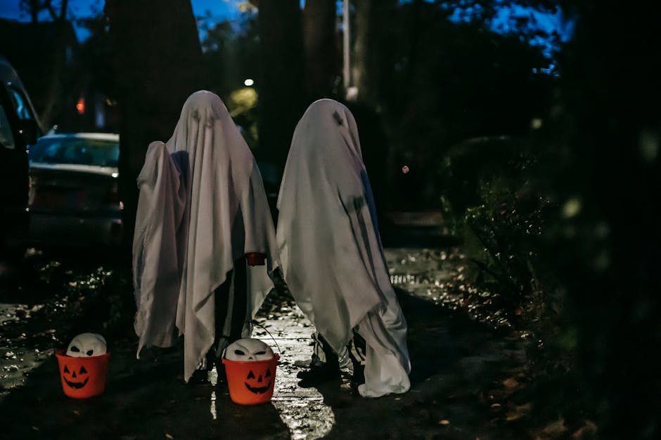 Two children dressed as ghosts in white sheets stand on a dimly lit path at night, holding orange pumpkin buckets with ghost faces. Trees and a parked car are visible in the background amidst the dark setting.