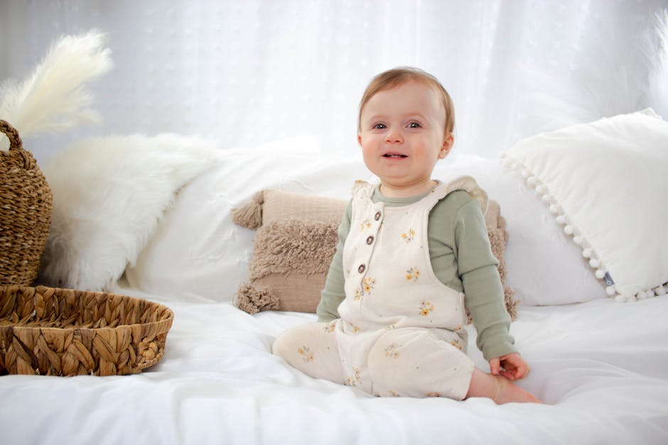 A baby sits on a white bed, wearing a floral-patterned onesie and green long-sleeve shirt. The room is decorated with fluffy pillows and a wicker basket, creating a cozy atmosphere.