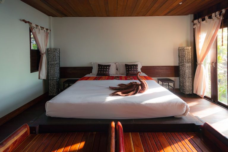 A cozy bedroom with a large bed adorned with a white duvet and red-orange accents. Two pillows rest at the head, and a brown towel is artistically folded in the center. Warm natural light filters through sheer curtains, illuminating the space.