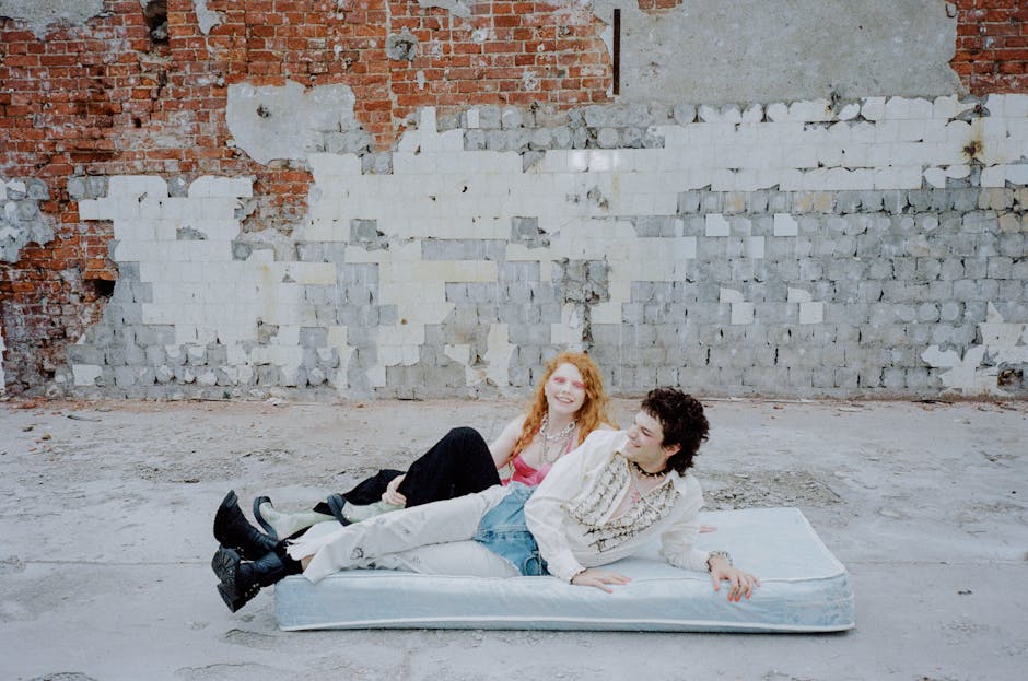 Two people, one with long red hair and the other with short dark hair, are lying on a mattress placed on rough concrete ground against a weathered brick and plaster wall. They appear relaxed and are smiling at each other.