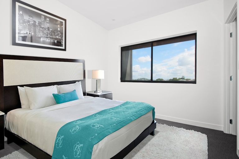 A modern bedroom with a large bed featuring white and turquoise bedding. A black and white cityscape photo hangs above the bed. A window to the right offers a view of the blue sky and trees. A bedside table with a lamp is on the left.