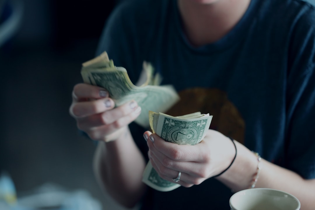 A person wearing a blue shirt is holding and counting multiple one-dollar bills. Their face is not visible in the image. A cup is positioned near the bottom right corner. The background is blurred.