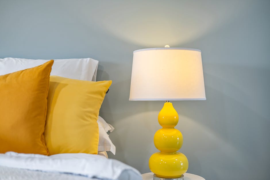 A cozy bedroom corner featuring a bed with white sheets and vibrant yellow pillows. Beside the bed is a stylish yellow lamp with a white lampshade, set against a light gray wall.