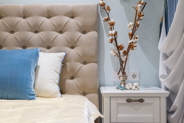 A neatly made bed with beige and blue pillows is next to a white nightstand with a drawer. The nightstand holds a decorative vase with cotton stems. The room has light blue walls and a tufted beige headboard. White curtains hang on the side.